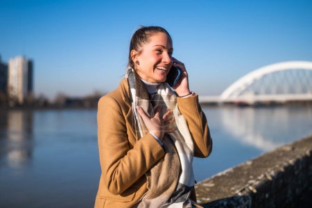 Photo for Surprised woman talking on a mobile phone  in warm clothing  and resting  by the river on a sunny winter day. - Royalty Free Image