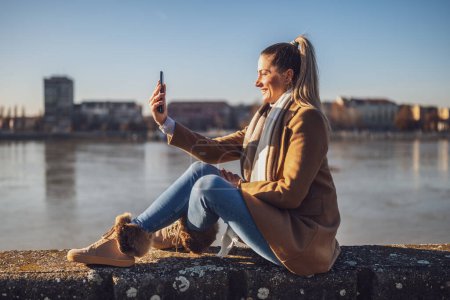 Foto de Hermosa mujer en ropa de abrigo tomando selfie con teléfono y disfruta de descansar junto al río en un día soleado de invierno. Imagen tonificada. - Imagen libre de derechos