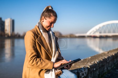 Photo for Beautiful woman in warm clothing using digital tablet and enjoys resting by the river on a sunny winter day. - Royalty Free Image
