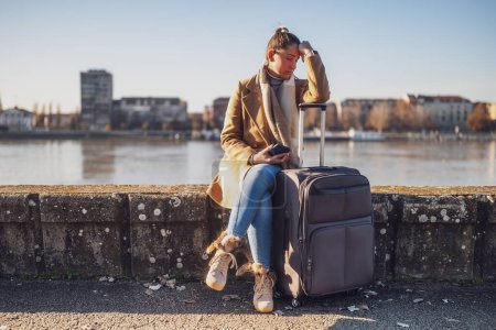 Téléchargez les photos : Femme touriste inquiète avec valise et téléphone perdu dans une ville assise au bord de la rivière et pensant. Image tonique. - en image libre de droit