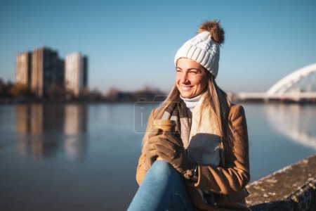 Téléchargez les photos : Belle femme en vêtements chauds aime boire du café et se reposer au bord de la rivière par une journée d'hiver ensoleillée. Image tonique. - en image libre de droit