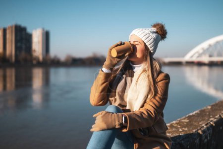 Foto de Hermosa mujer en ropa de abrigo disfruta de beber café y descansar junto al río en un día soleado de invierno. Imagen tonificada. - Imagen libre de derechos