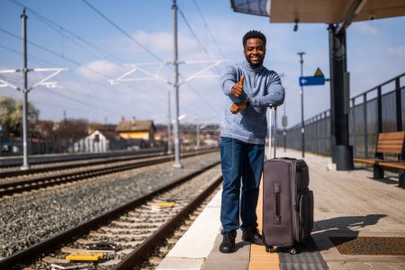 Téléchargez les photos : Homme heureux avec valise debout sur la gare et montrant pouce vers le haut. - en image libre de droit