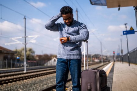 Téléchargez les photos : Homme inquiet regardant son horloge debout avec une valise sur une gare. - en image libre de droit