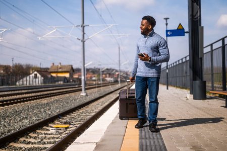 Foto de Hombre feliz con una maleta usando el teléfono mientras camina en la estación de tren. - Imagen libre de derechos