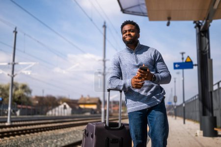 Téléchargez les photos : Homme en colère avec une valise et un téléphone portable debout sur une gare. - en image libre de droit