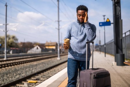 Téléchargez les photos : Un homme fatigué buvant du café debout avec une valise sur une gare. - en image libre de droit