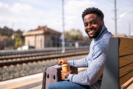 Foto de Hombre feliz con maleta disfruta bebiendo café mientras está sentado en un banco en la estación de tren. - Imagen libre de derechos