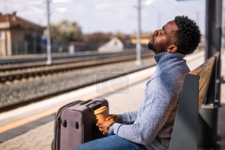 Téléchargez les photos : Homme fatigué buvant du café assis sur le banc avec valise sur une gare. - en image libre de droit
