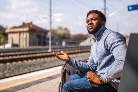 Téléchargez les photos : Homme en colère avec une valise et un téléphone portable assis sur un banc à la gare. - en image libre de droit