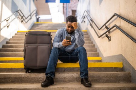 Téléchargez les photos : Homme inquiet avec un téléphone et une valise assis sur un escalier à la gare. - en image libre de droit