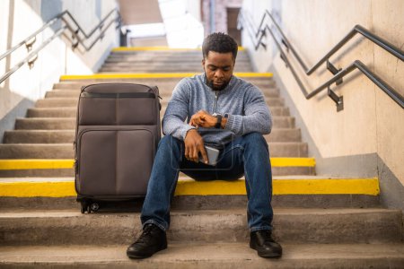 Téléchargez les photos : Homme inquiet regardant l'horloge avec un téléphone et une valise tout en étant assis sur un escalier à la gare. - en image libre de droit
