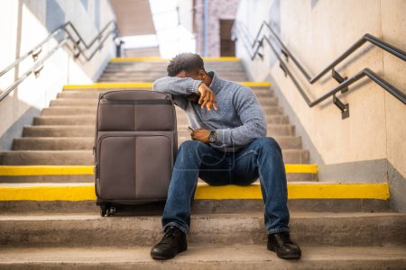 Foto de Hombre preocupado con un teléfono y una maleta sentado en una escalera en la estación de tren. - Imagen libre de derechos