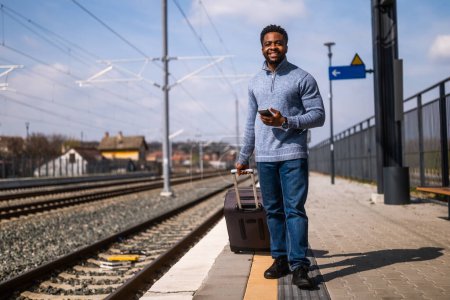 Téléchargez les photos : Homme heureux avec une valise en utilisant le téléphone tout en marchant sur la gare. - en image libre de droit