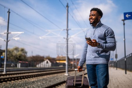 Foto de Hombre feliz con una maleta usando el teléfono mientras está de pie en la estación de tren. - Imagen libre de derechos