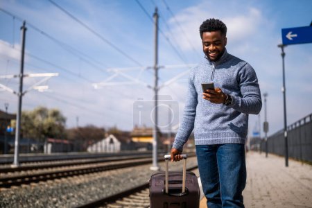 Photo for Happy man with a suitcase using  phone while standing on the railway station. - Royalty Free Image