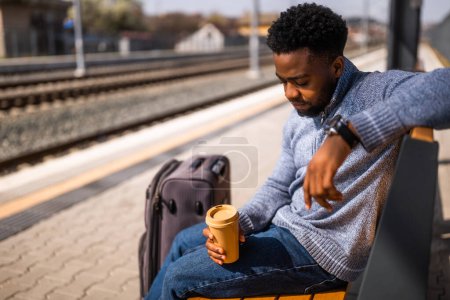 Foto de Hombre cansado bebiendo café mientras está sentado en el banco con la maleta en una estación de tren. - Imagen libre de derechos
