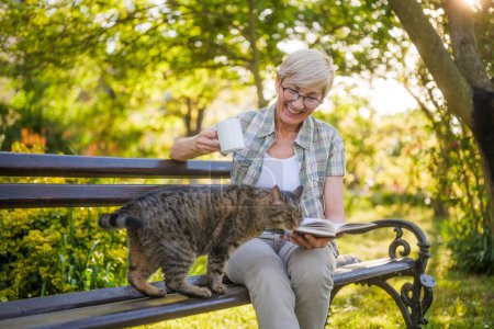 Photo for Happy senior woman enjoys reading book ,drinking coffee  and spending time with her cat on a bench in  garden. - Royalty Free Image