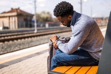 Téléchargez les photos : Homme inquiet regardant son horloge assis sur un banc à la gare. - en image libre de droit