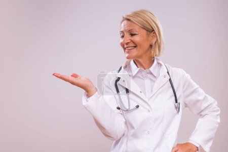 Photo for Portrait of mature female doctor showing palm of hand on gray background. - Royalty Free Image