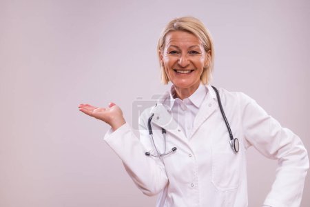 Photo for Portrait of mature female doctor showing palm of hand on gray background. - Royalty Free Image