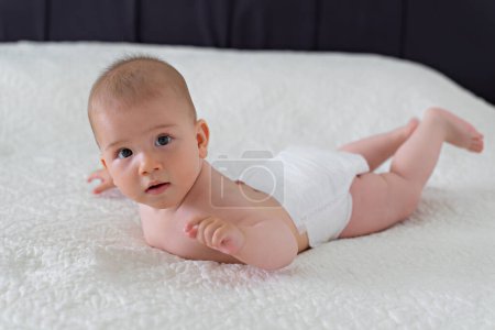 Photo for Little cute 6 month old baby in a diaper lies on his stomach on a white blanket - Royalty Free Image