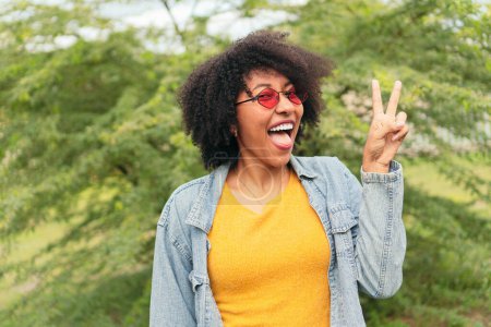Photo for Afro woman making peace sign with fingers while posing over a natural green background - Royalty Free Image