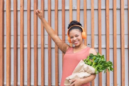 Radiant young woman raising her fist in triumph, wearing orange headphones, holding fresh greens in a paper bag.