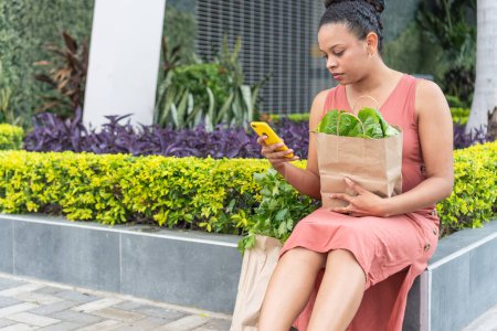 Young woman browsing her smartphone after buying fresh vegetables in an urban setting.