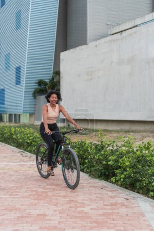 A woman with a joyful expression rides a bicycle on a paved pathway, with modern buildings in the background.