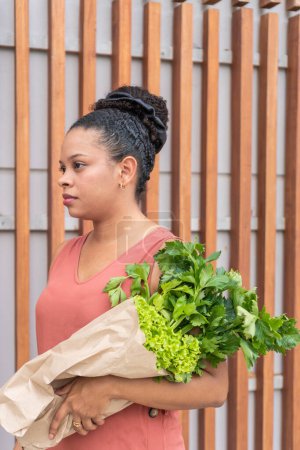 Pensive woman holding a bag of leafy vegetables against a wooden slat backdrop.