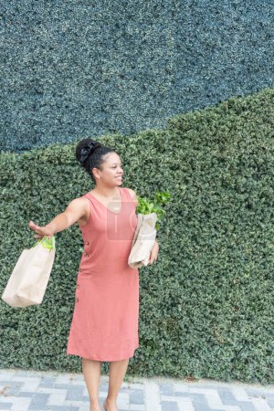 Photo for Cheerful young woman in a coral dress holding paper bags of fresh produce with a textured hedge in the background. - Royalty Free Image