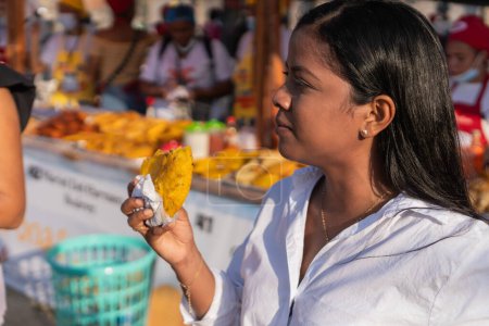 Téléchargez les photos : Femme dégustant une collation frite fraîche dans un marché de nourriture de rue animé. - en image libre de droit