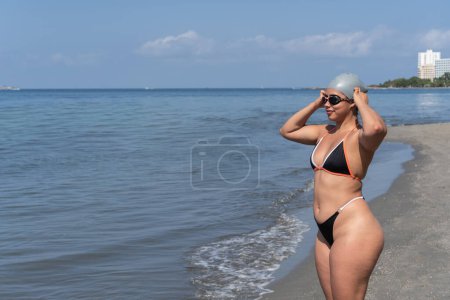 A swimmer stands by the ocean, adjusting her swim cap, ready to dive in.