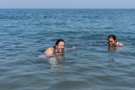 Two women smiling while swimming in clear blue sea.
