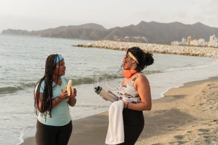 Zwei Frauen in Trainingsanzügen machen am Strand eine Snack-Pause, wobei die eine eine Banane isst und die andere eine Wasserflasche in der Hand hält.