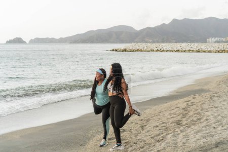 Deux femmes profitant d'un étirement de jambe sur une plage calme, avec des collines en arrière-plan.
