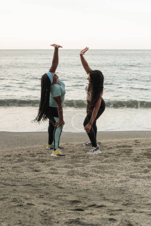 Two women enjoy a dynamic stretching exercise on the beach at twilight.