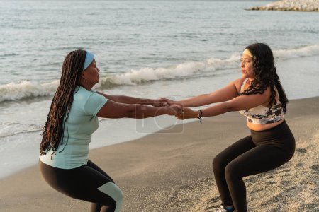 A pair of women in workout attire hold hands while performing squats on a sandy beach, with waves and rocks in the background.