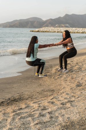 Workout partners engage in squats, assisting each other on a sunlit beach with coastal mountains in the background.