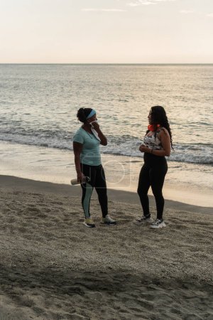 Zwei Frauen unterhalten sich nach dem Workout an einem ruhigen Strand in der Abenddämmerung.