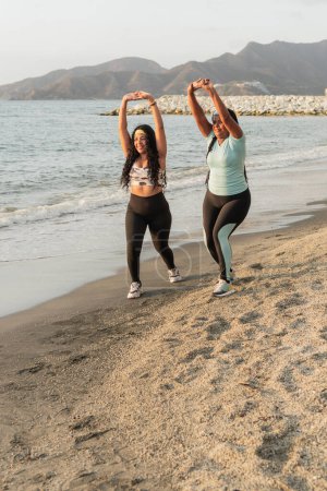 Two women with raised arms in a celebratory gesture during their beach workout at sunset.