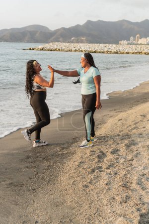 Zwei fröhliche High-Five-Frauen beim Beach-Workout, die ihre Fitness-Fortschritte feiern.