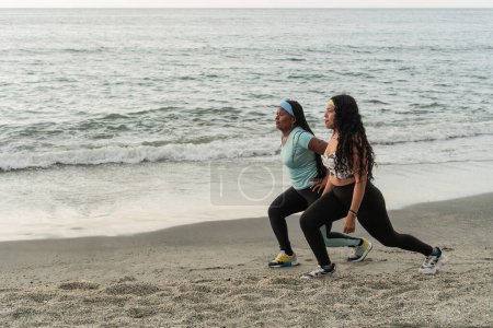 Women in workout gear perform lunges on a sandy beach with gentle waves in the background.