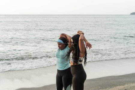 Workout partners perform synchronized arm stretches by the sea.