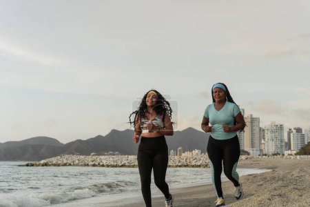 Smiling women running on a sandy beach with a cityscape and mountains in the distance.