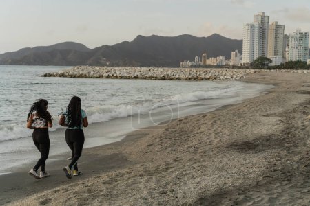 Two runners enjoy a scenic jog on the beach with cityscape and mountains in the distance.