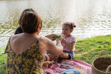 Lächelndes Kind stößt mit Familie beim Picknick am schimmernden See an.