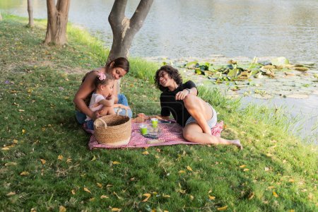 Una familia se ríe y juega mientras descansa en una manta de picnic junto a un lago pintoresco.