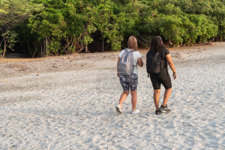 Dos amigos con mochilas caminan lado a lado en una playa de arena con un telón de fondo de exuberantes árboles verdes.